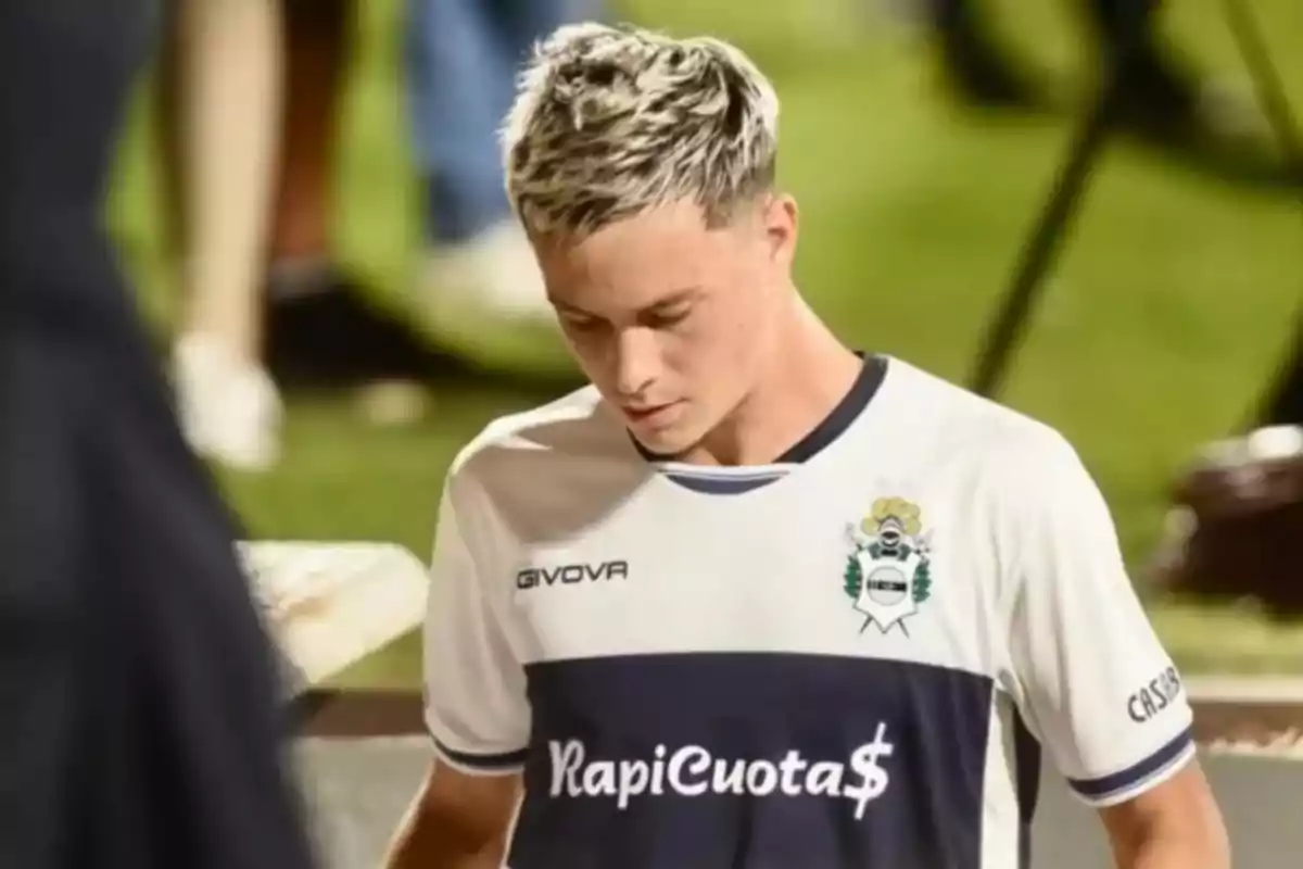 Un jugador de fútbol con cabello rubio y camiseta blanca con detalles azules camina en el campo.