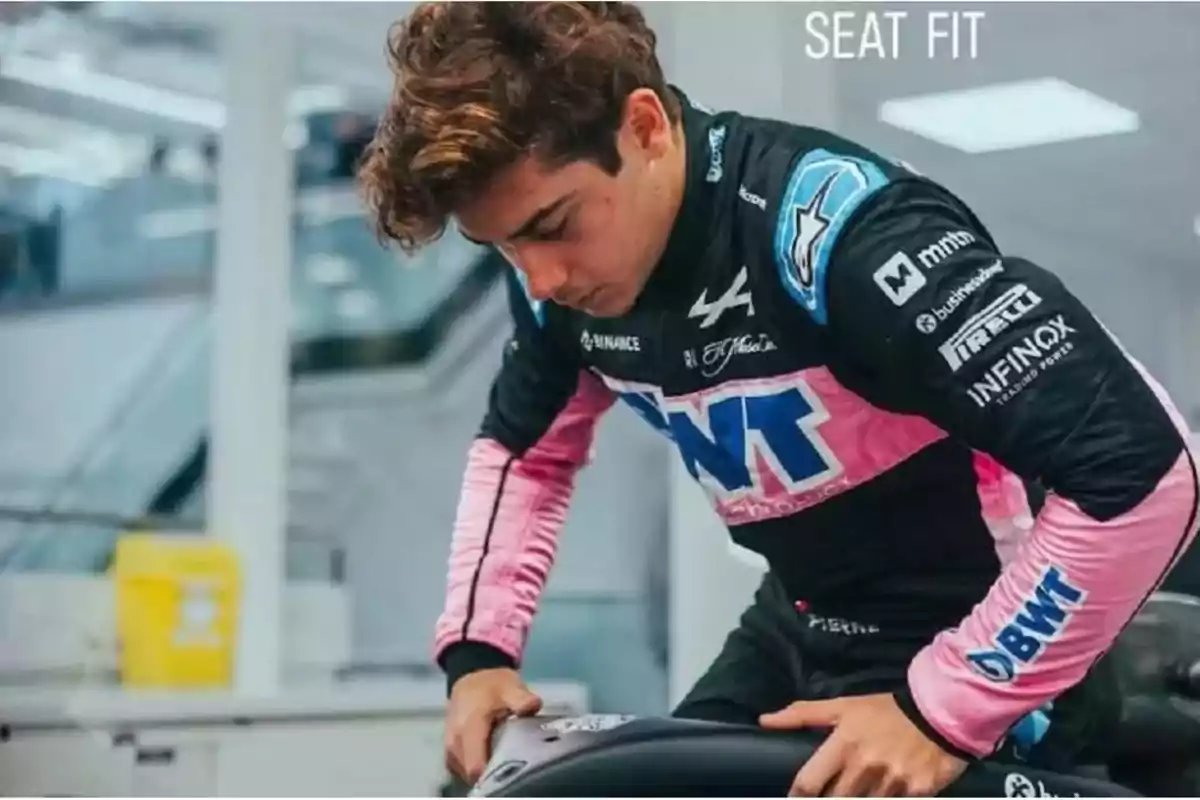 Un piloto de carreras ajustando su asiento en un entorno de taller, vistiendo un traje de carreras con detalles en rosa y azul.