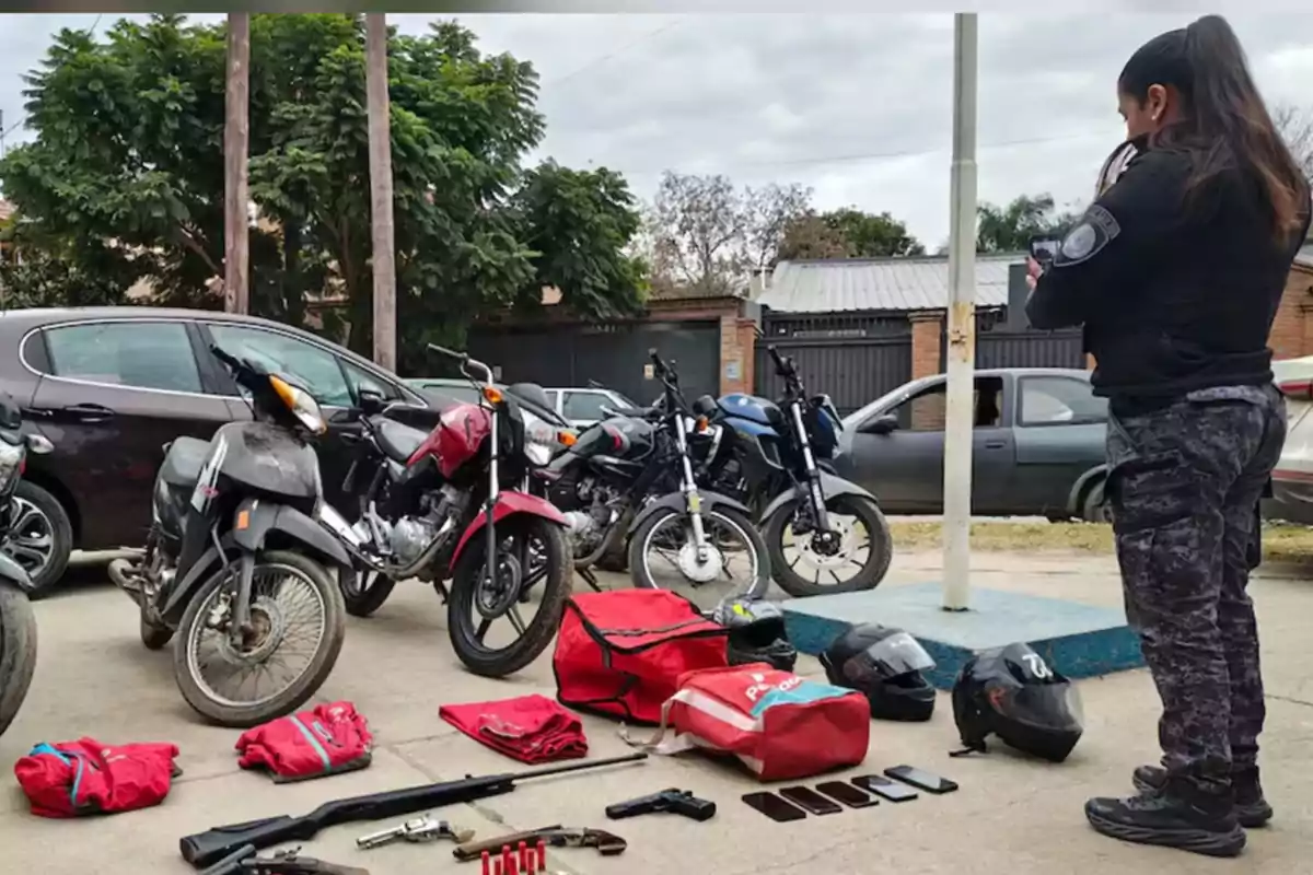 Una mujer policía observa varias motocicletas estacionadas junto a autos y una serie de objetos incautados en el suelo, incluyendo cascos, mochilas rojas, armas, municiones y teléfonos móviles Una mujer policía observa varias motocicletas estacionadas junto a autos y una serie de objetos incautados en el suelo, incluyendo cascos, mochilas rojas, armas, municiones y teléfonos móviles