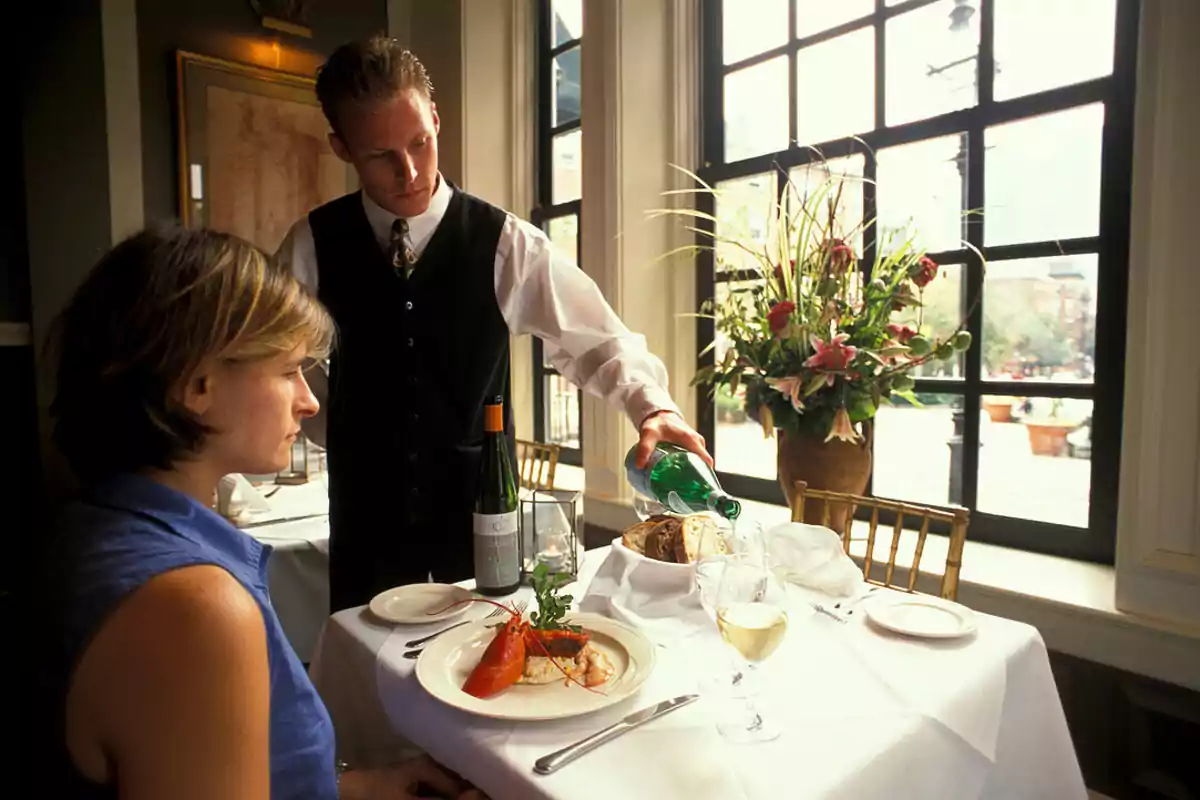 Una persona sentada en un restaurante mientras un camarero sirve agua en un vaso sobre una mesa elegantemente puesta con un plato de comida y una botella de vino. Una persona sentada en un restaurante mientras un camarero sirve agua en un vaso sobre una mesa elegantemente puesta con un plato de comida y una botella de vino.