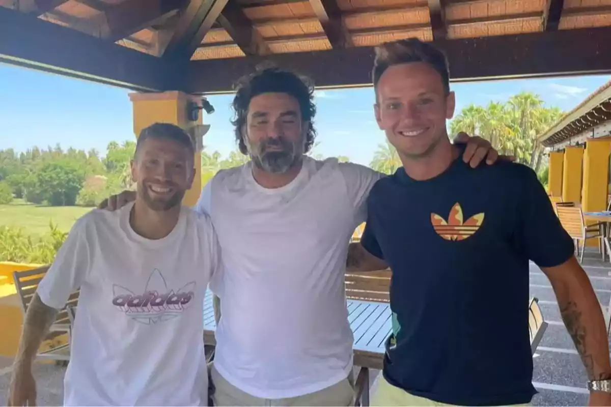 Three men smile and pose together under a wooden roof in an outdoor setting with greenery and tables around. Three men smile and pose together under a wooden roof in an outdoor setting with greenery and tables around.