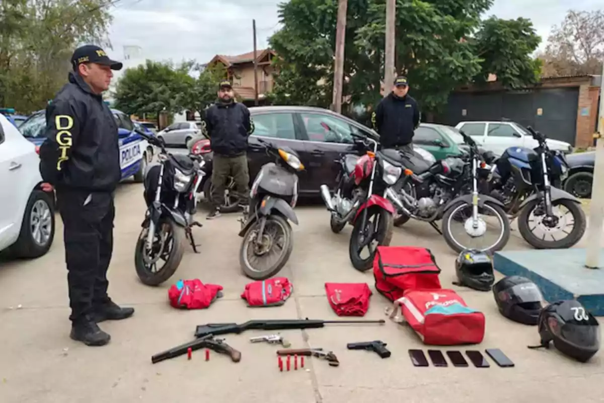 Tres policías de pie junto a varias motocicletas, armas, cascos, mochilas de reparto y teléfonos móviles incautados, todo dispuesto en el suelo frente a ellos en un operativo policial.