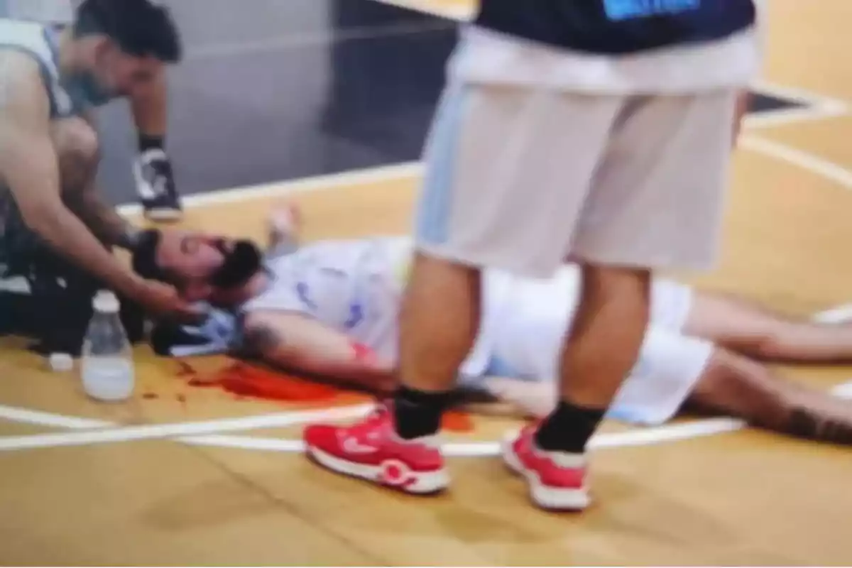 Un jugador de baloncesto está tendido en el suelo con una herida en la cabeza mientras otro jugador lo asiste en la cancha.