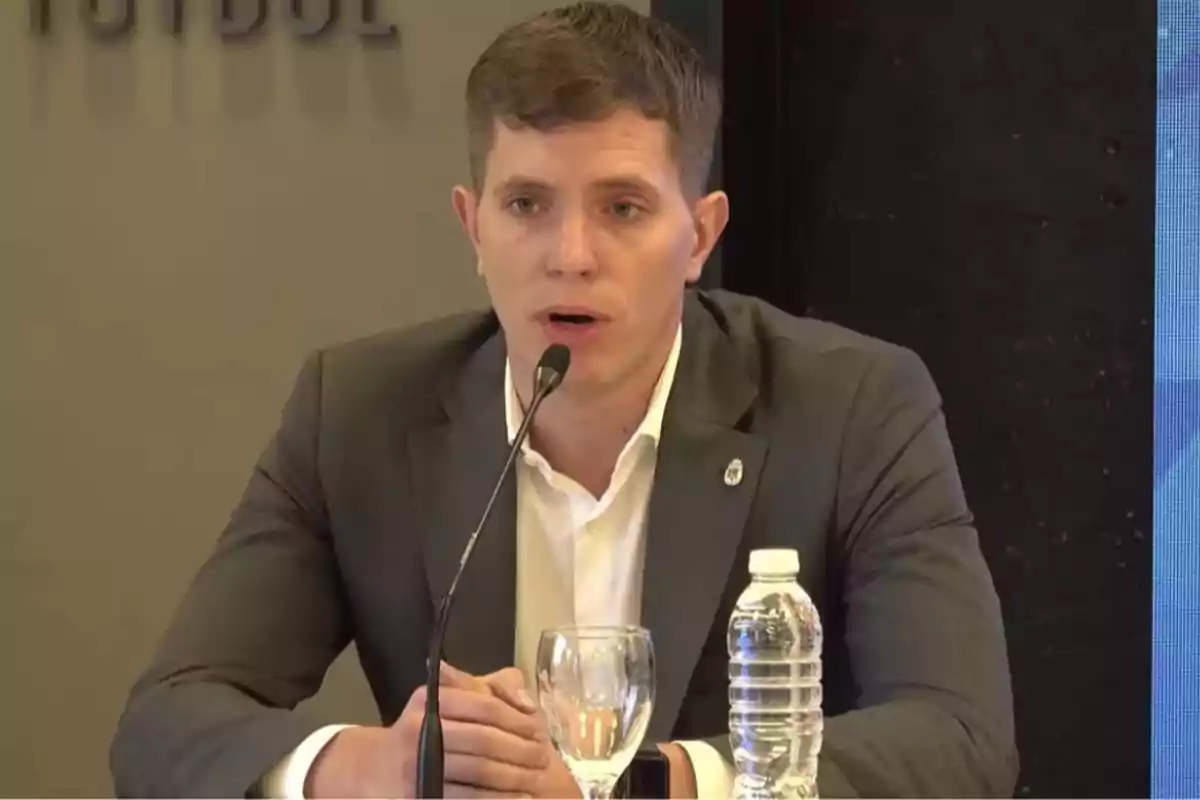 Hombre joven con saco gris y camisa blanca hablando frente a un micrófono en una mesa con una botella de agua y una copa de vidrio Hombre joven con saco gris y camisa blanca hablando frente a un micrófono en una mesa con una botella de agua y una copa de vidrio