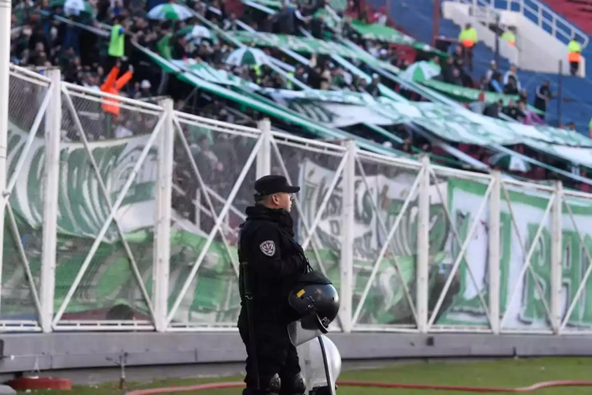 A police officer in riot gear stands guard in front of a grandstand full of fans with green flags and umbrellas at a soccer stadium.