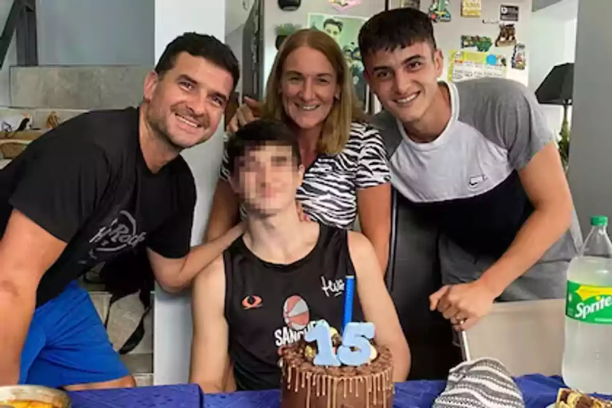 Four people smile around a table with a chocolate birthday cake that has the number 15 and a blue candle; in the background, there is a refrigerator with magnets and a bottle of Sprite on the table. Four people smile around a table with a chocolate birthday cake that has the number 15 and a blue candle; in the background, there is a refrigerator with magnets and a bottle of Sprite on the table.