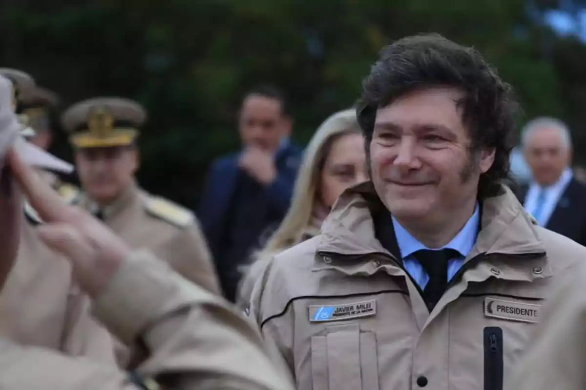 Un hombre con chaqueta beige y corbata azul oscuro sonríe mientras es saludado por un militar, con otras personas uniformadas en el fondo.