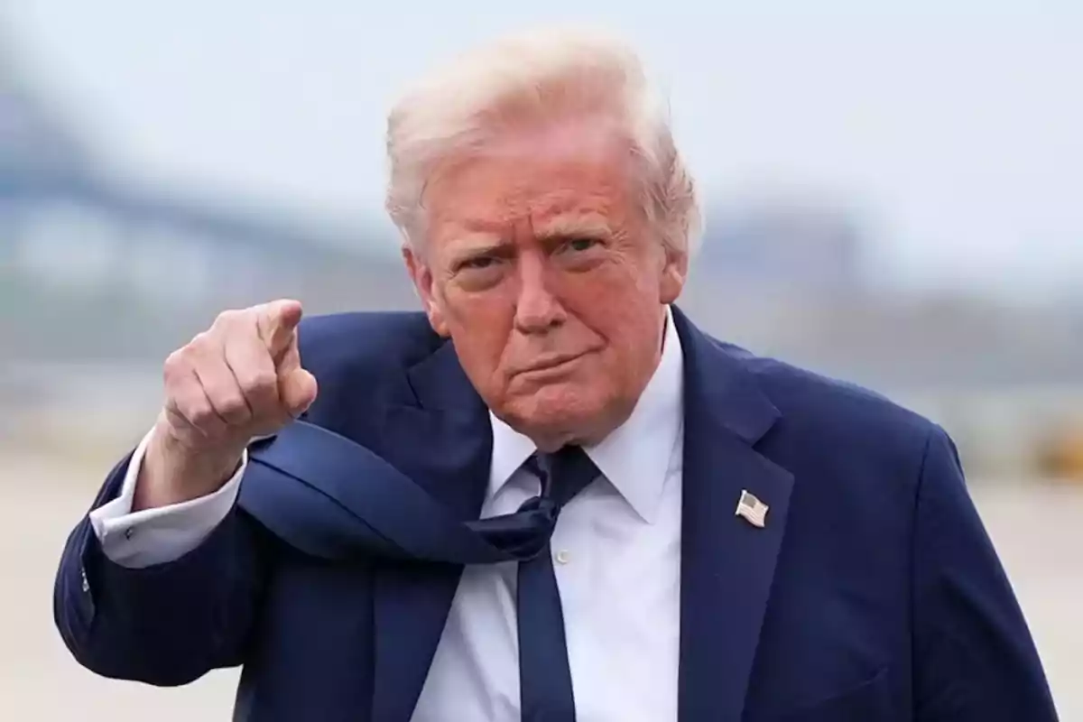 A light-haired man in a dark suit points forward with a serious expression.
