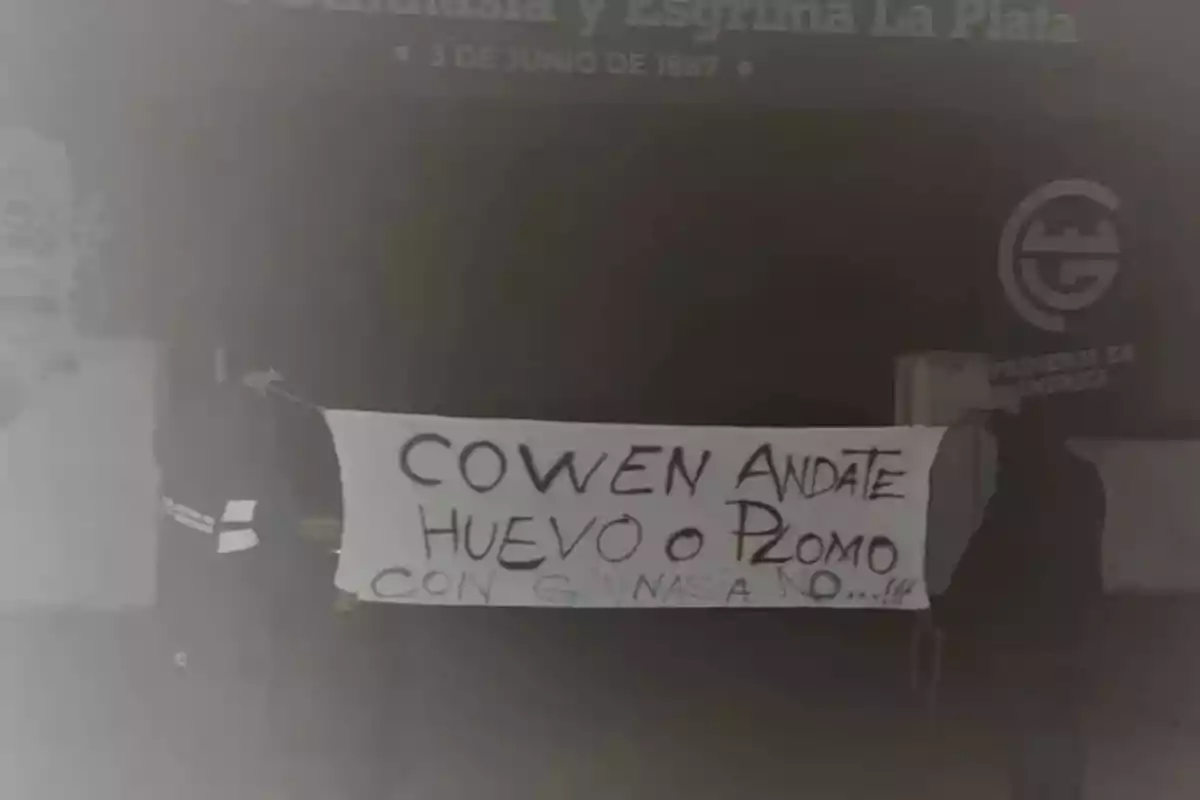 Dos personas sostienen una pancarta con un mensaje escrito a mano frente a un edificio que parece ser un club deportivo. Dos personas sostienen una pancarta con un mensaje escrito a mano frente a un edificio que parece ser un club deportivo.