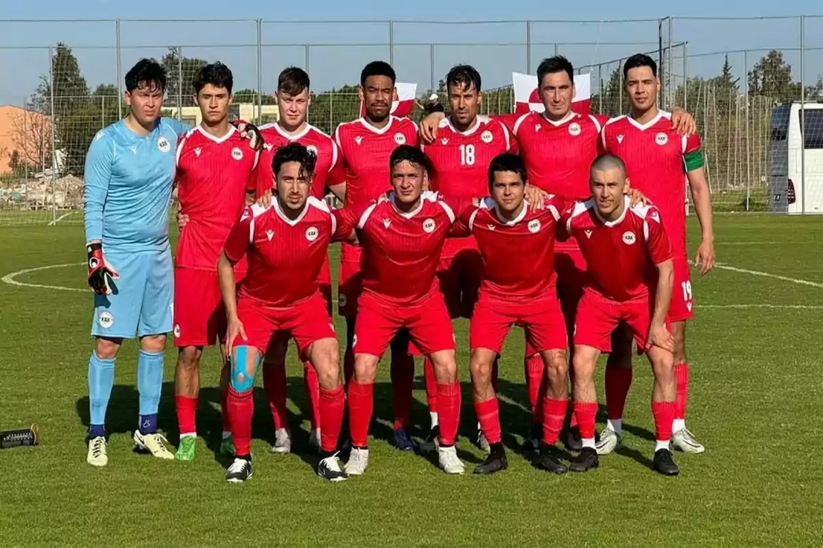 Once jugadores de fútbol posan en el campo antes de un partido, todos con uniforme rojo excepto el portero que viste de azul, con una portería y árboles al fondo.