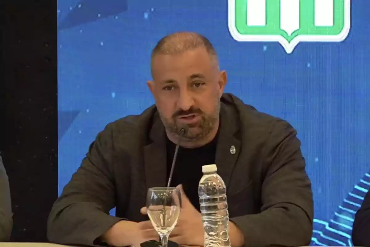 Hombre de barba y cabello corto hablando en una conferencia con un micrófono frente a él y una botella de agua y una copa sobre la mesa Hombre de barba y cabello corto hablando en una conferencia con un micrófono frente a él y una botella de agua y una copa sobre la mesa