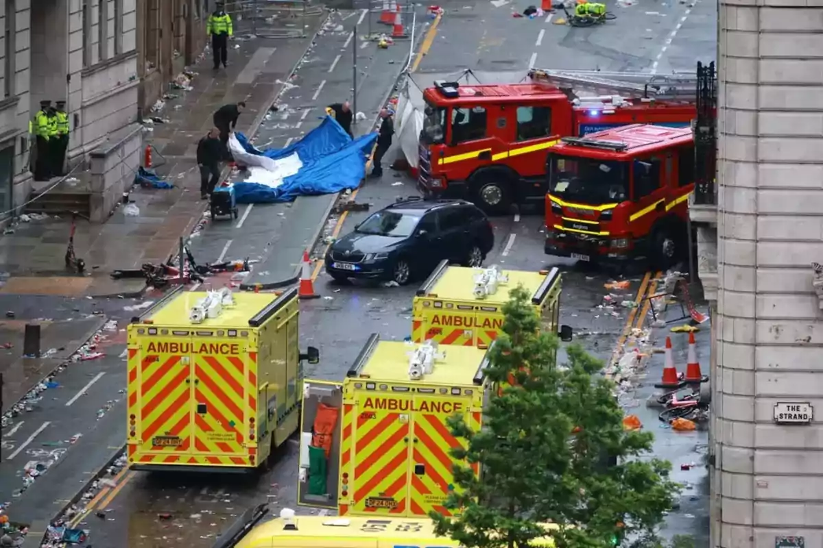 Varios vehículos de emergencia, incluyendo ambulancias y camiones de bomberos, están atendiendo una situación en una calle llena de escombros y basura, mientras algunos policías y personal de emergencia trabajan en la escena.
