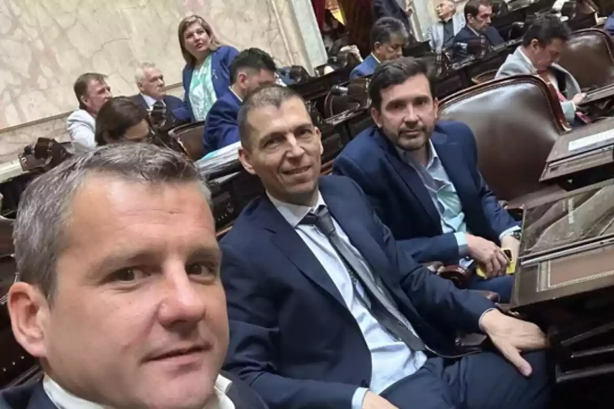 Un grupo de personas en trajes formales sentados en un salón con escritorios de madera.