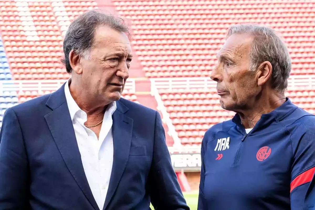 Dos hombres conversan en las gradas de un estadio de fútbol uno lleva traje azul y el otro viste ropa deportiva azul con un escudo rojo Dos hombres conversan en las gradas de un estadio de fútbol uno lleva traje azul y el otro viste ropa deportiva azul con un escudo rojo