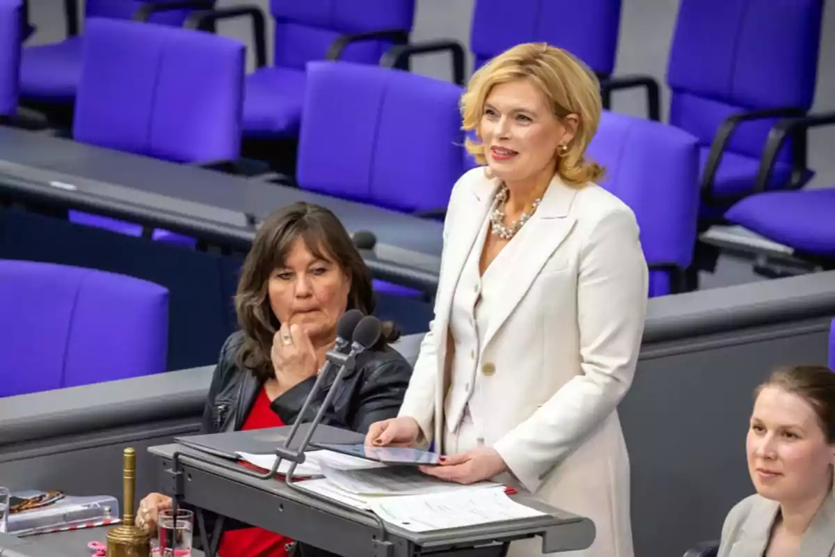 Una mujer de pie en un podio hablando mientras otras dos mujeres están sentadas cerca en un entorno de asamblea con asientos morados. Una mujer de pie en un podio hablando mientras otras dos mujeres están sentadas cerca en un entorno de asamblea con asientos morados.