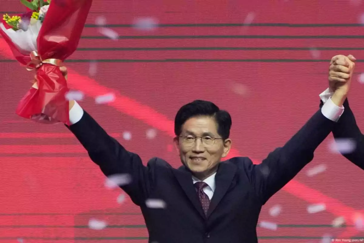 Hombre con gafas y traje oscuro sonríe mientras sostiene un ramo de flores y levanta la mano de otra persona en un escenario con fondo rojo y confeti en el aire Hombre con gafas y traje oscuro sonríe mientras sostiene un ramo de flores y levanta la mano de otra persona en un escenario con fondo rojo y confeti en el aire