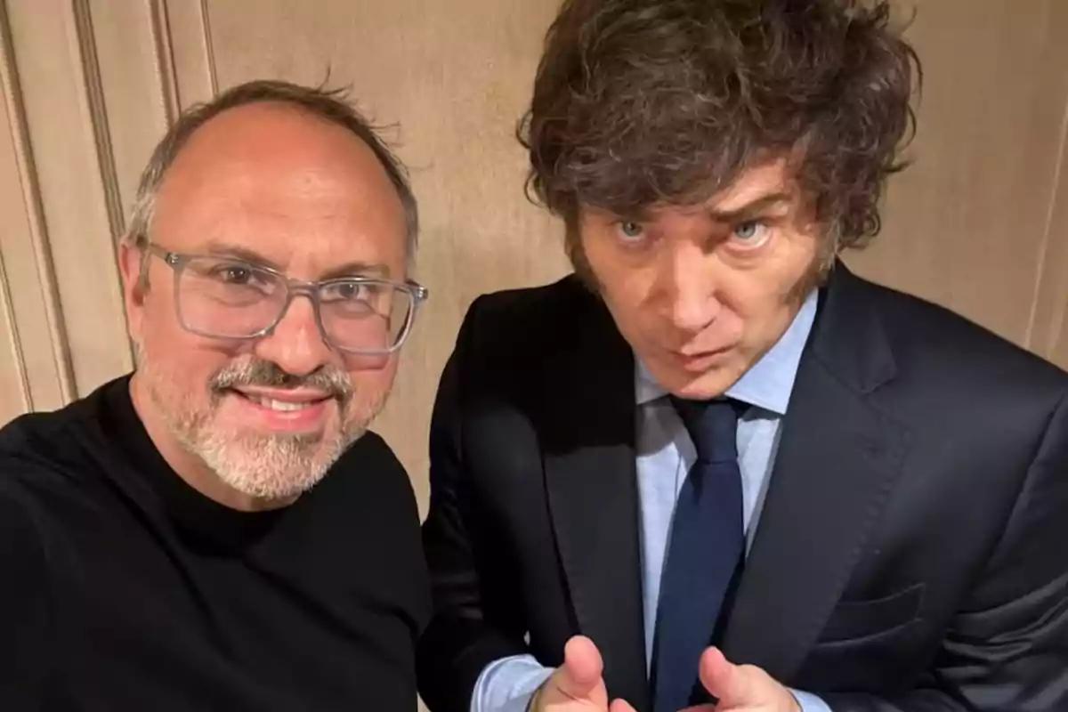 Two men pose together for a selfie; one of them is wearing glasses and a black t-jersey, while the other is dressed in a dark suit and blue tie.