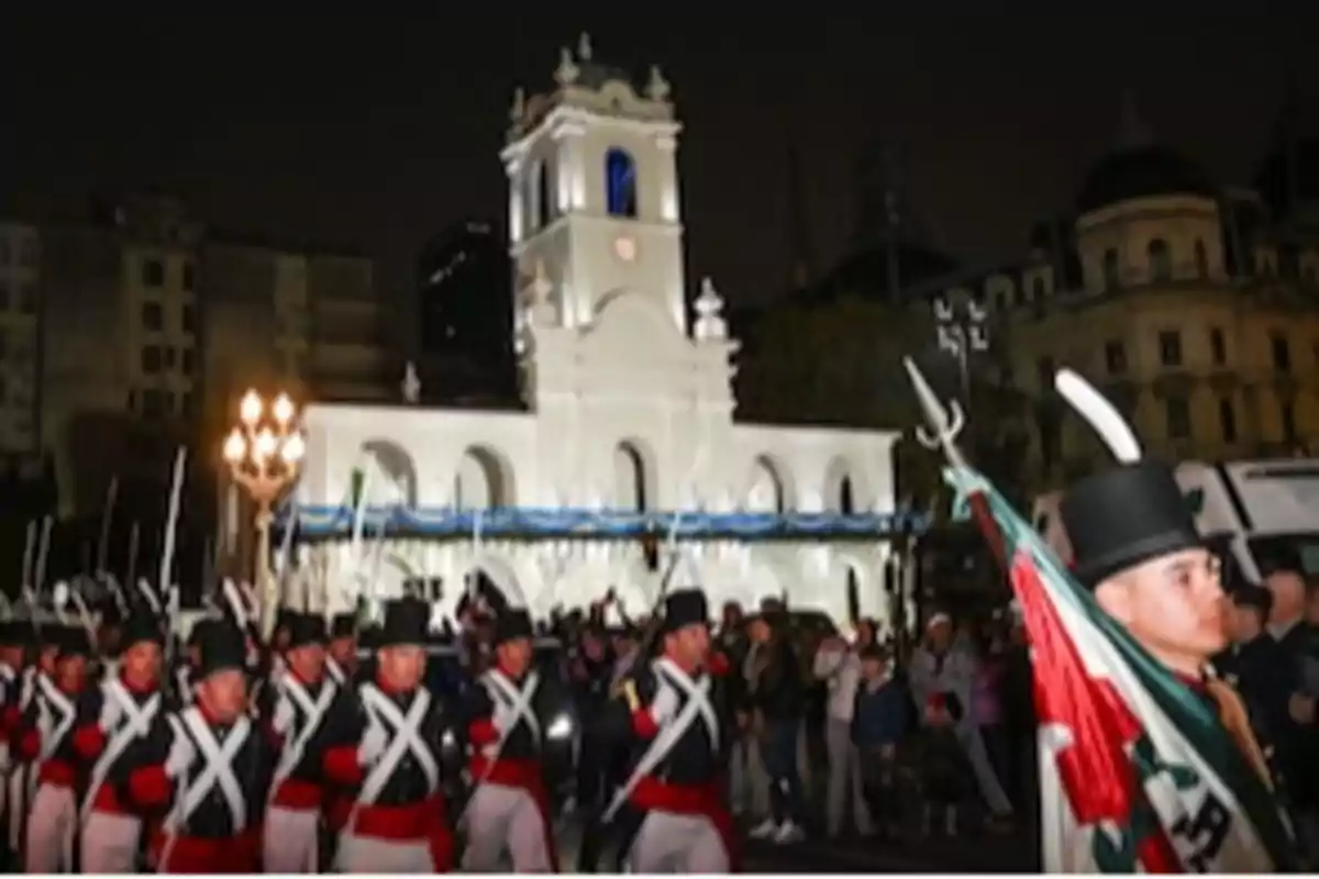 A group of soldiers in historical uniforms marches in front of a building lit up at night, while a crowd watches. A group of soldiers in historical uniforms marches in front of a building lit up at night, while a crowd watches.