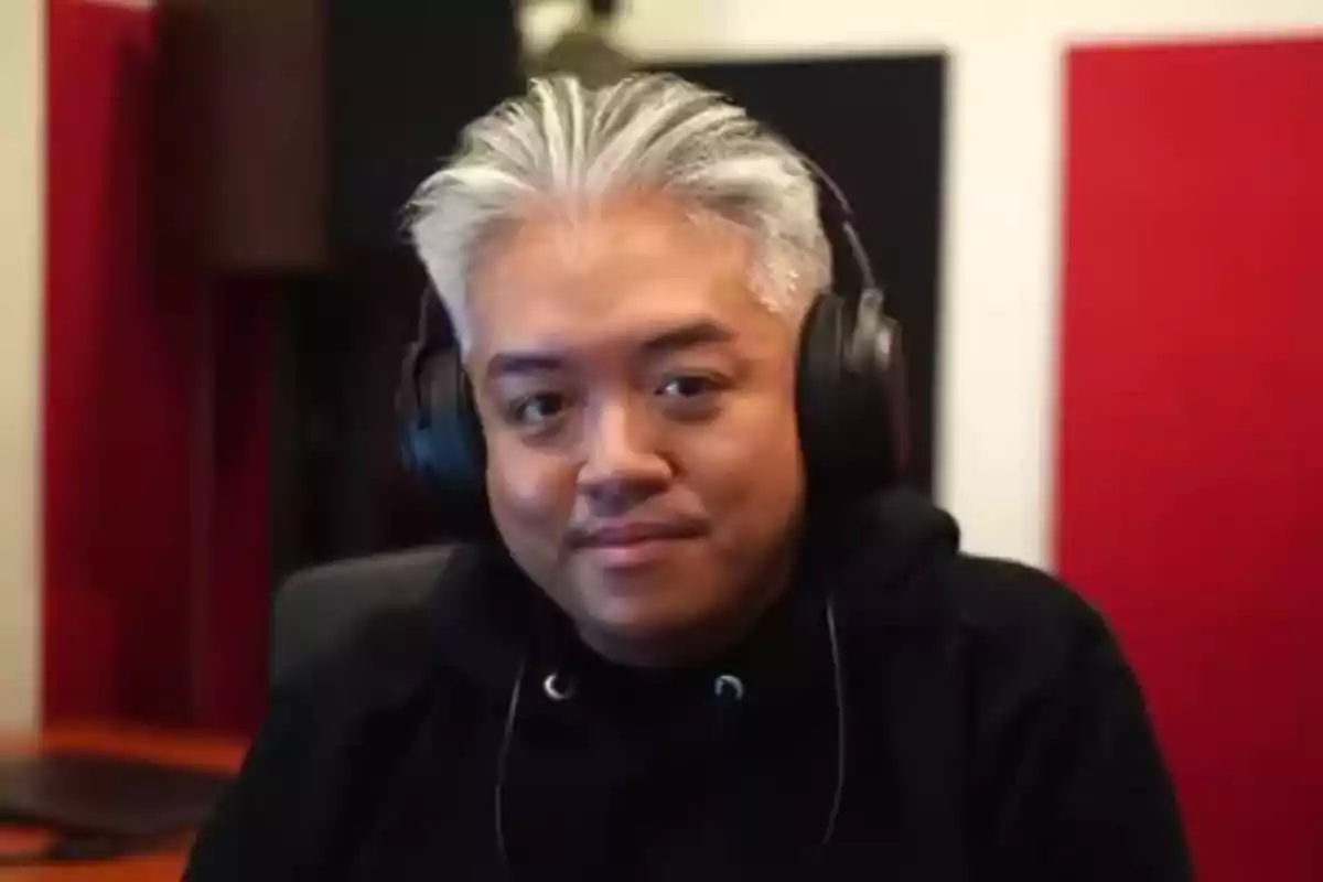 Gray-haired man wearing black headphones and a dark sweatjersey sitting in a room with red and black acoustic panels. Gray-haired man wearing black headphones and a dark sweatjersey sitting in a room with red and black acoustic panels.