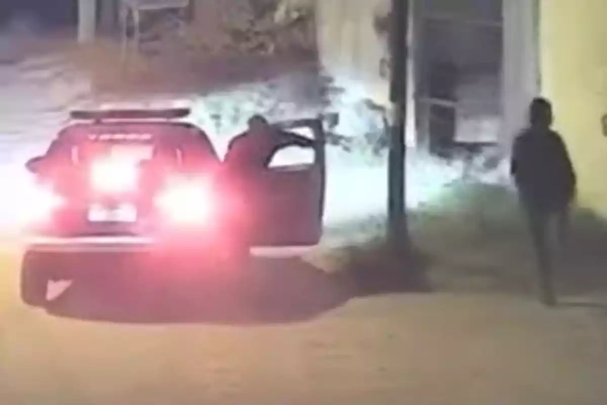 Una camioneta con las luces traseras encendidas se detiene en una calle de noche mientras una persona se baja del vehículo y otra camina cerca de la acera. Una camioneta con las luces traseras encendidas se detiene en una calle de noche mientras una persona se baja del vehículo y otra camina cerca de la acera.