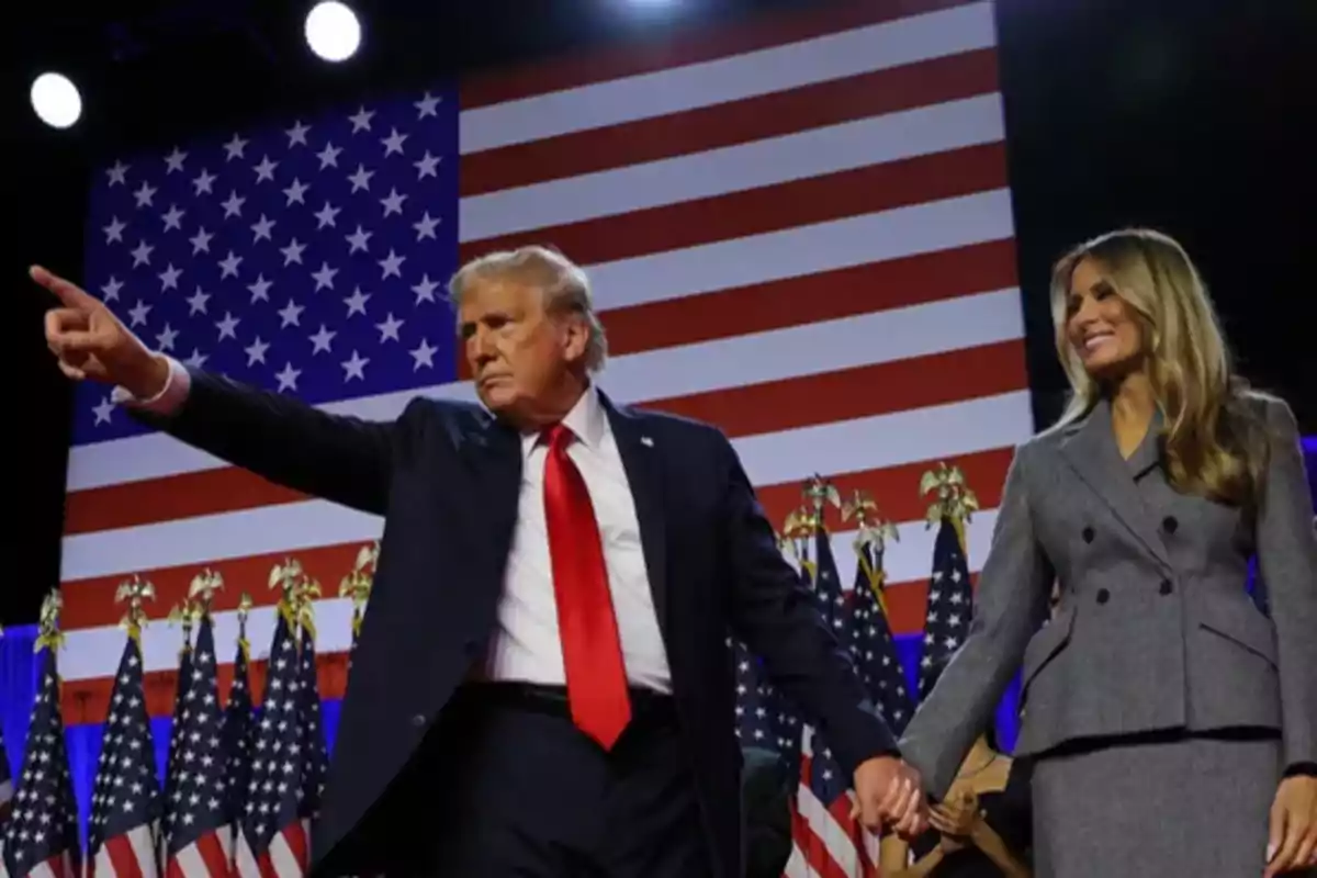 Dos personas de pie en un escenario con una bandera de Estados Unidos de fondo, una de ellas señala hacia adelante mientras sostienen las manos.