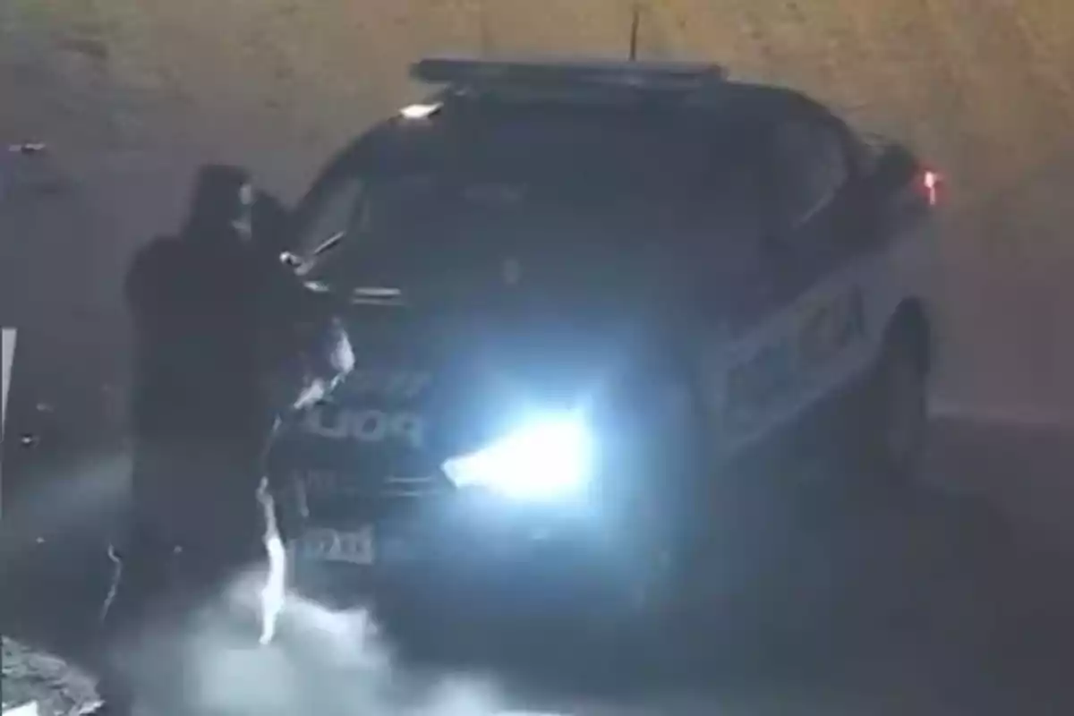 Persona encapuchada de pie frente a un auto de policía con las luces encendidas en la noche Persona encapuchada de pie frente a un auto de policía con las luces encendidas en la noche