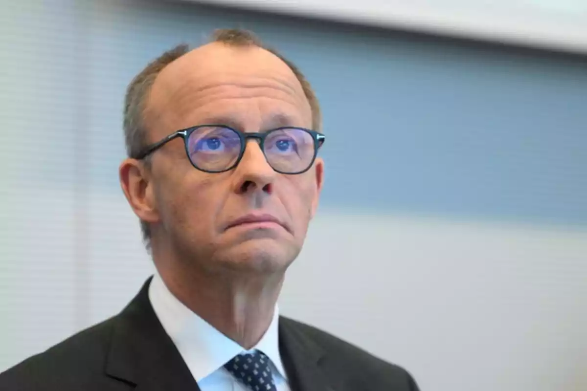Un hombre con gafas y traje oscuro mira hacia arriba con expresión seria en un entorno de oficina. Un hombre con gafas y traje oscuro mira hacia arriba con expresión seria en un entorno de oficina.