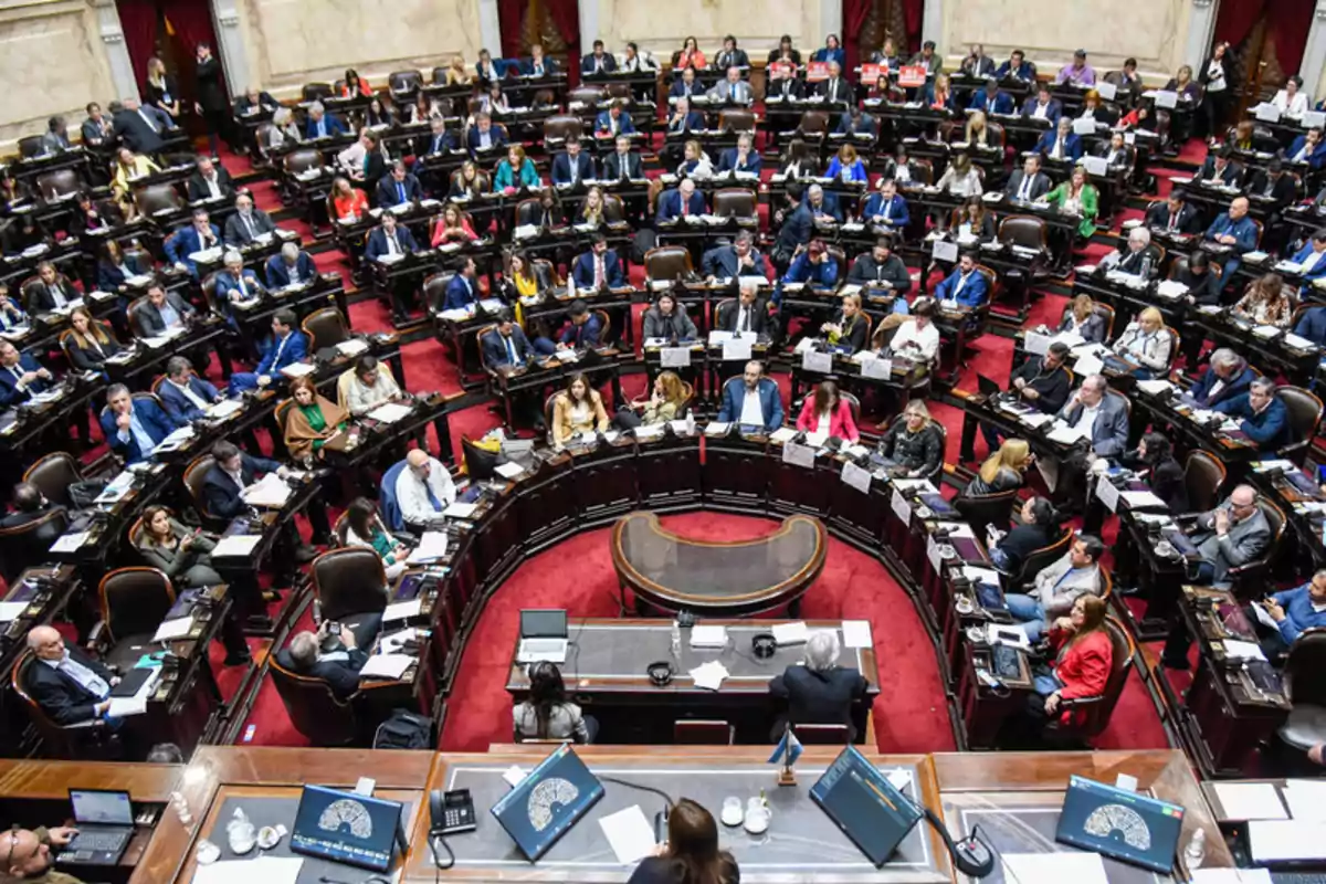 Vista aérea de una sesión parlamentaria con numerosos legisladores sentados en sus escaños en un recinto con alfombra roja y escritorios de madera.