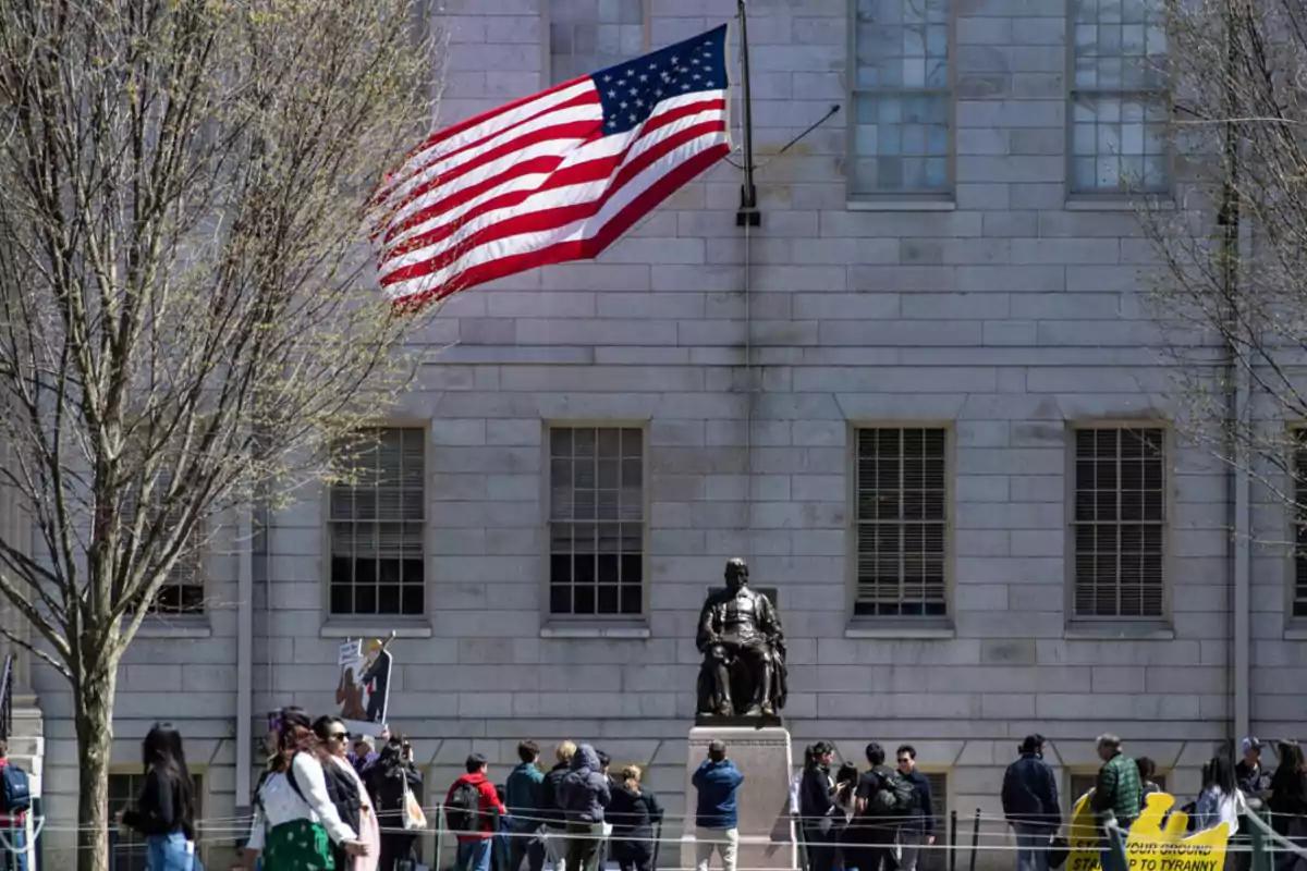 A group of people gathers around a statue in front of a stone building with a United States flag waving at the top.