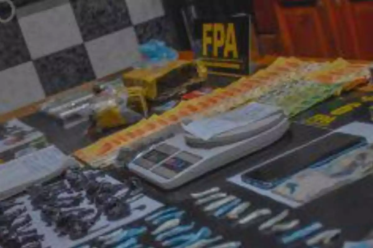 Objetos incautados sobre una mesa incluyendo una balanza, envoltorios, un teléfono móvil y un cartel con las siglas FPA en un entorno policial Objetos incautados sobre una mesa incluyendo una balanza, envoltorios, un teléfono móvil y un cartel con las siglas FPA en un entorno policial