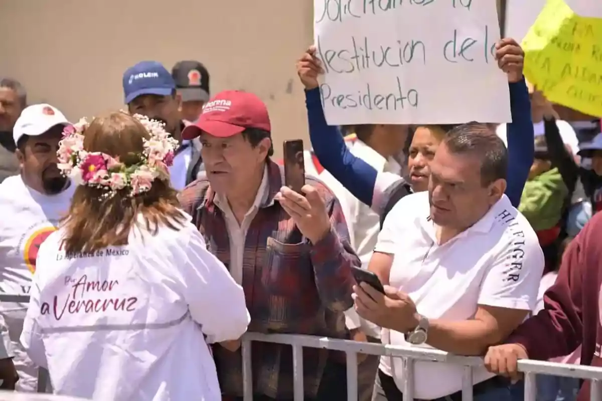 A group of people is gathered behind a fence, some holding signs and mobile phones, while a woman with a flower crown and a white jacket interacts with them.