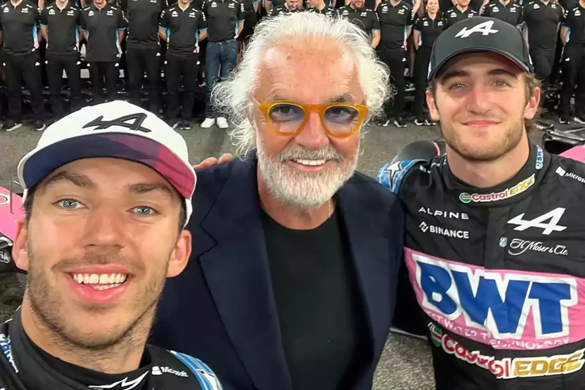 A group of people smiles for a photo, two of them wearing caps and racing uniforms, while the person in the center wears glasses and a dark jacket; in the background, a team of people dressed in black is lined up.