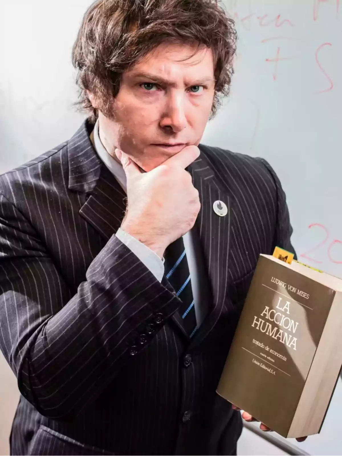 Hombre de cabello castaño con traje a rayas sosteniendo un libro titulado La Acción Humana y posando pensativo frente a un pizarrón Hombre de cabello castaño con traje a rayas sosteniendo un libro titulado La Acción Humana y posando pensativo frente a un pizarrón