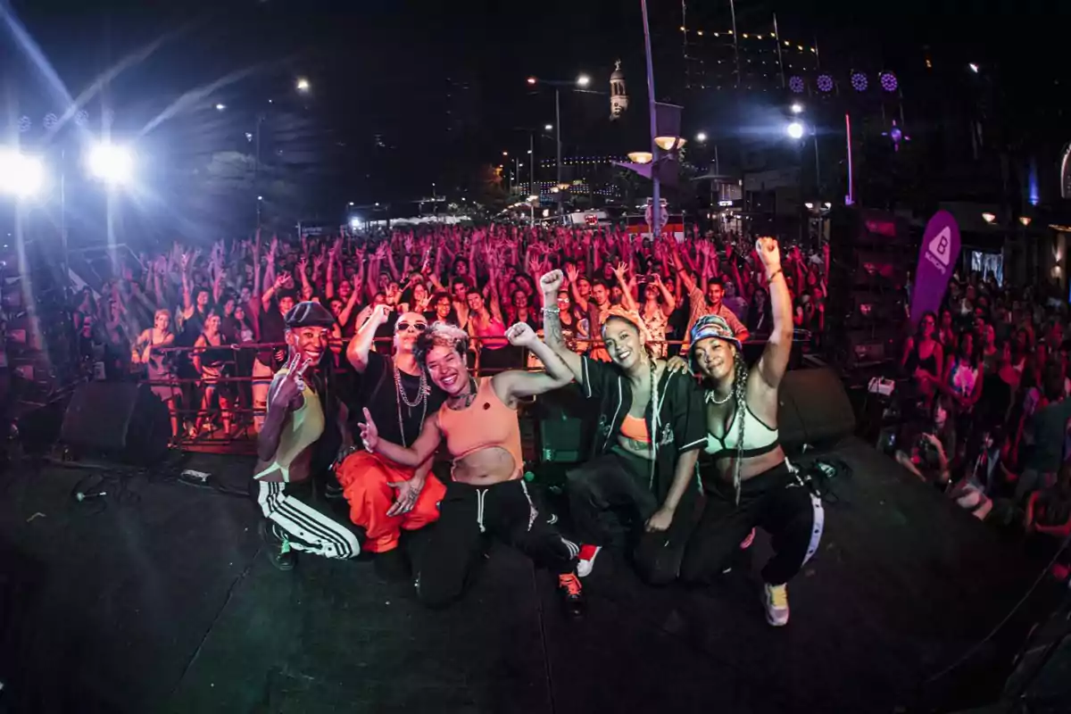 Five people pose smiling on stage in front of an excited crowd during an outdoor nighttime concert.