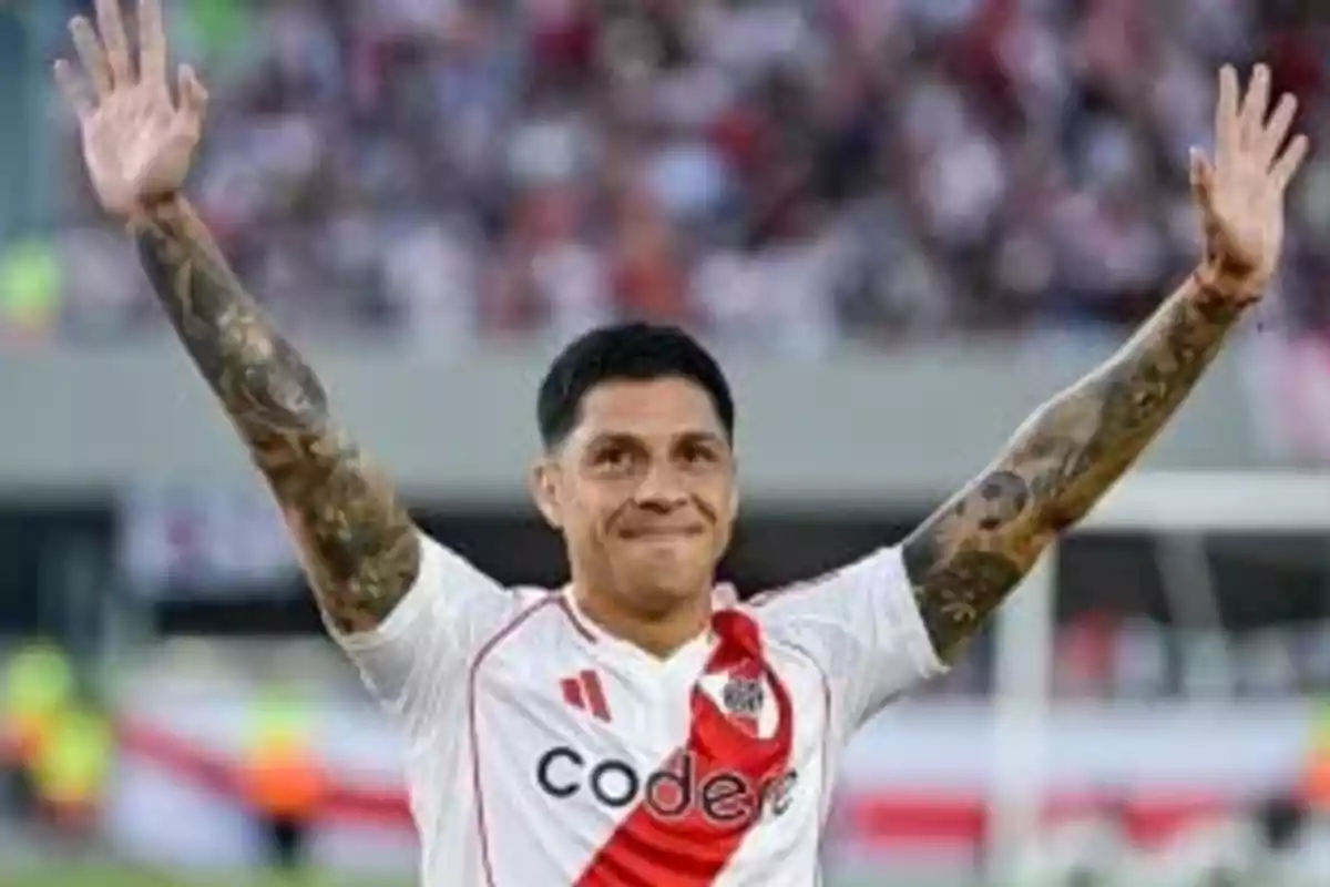 Un jugador de fútbol con uniforme blanco y rojo levanta los brazos en señal de celebración en un estadio.