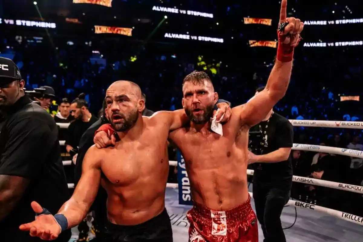 Dos luchadores en el ring, uno con el brazo alrededor del otro, celebrando después de un combate, con el público y las luces del estadio de fondo.