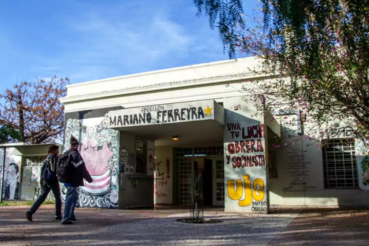 Dos personas caminan hacia la entrada de un edificio llamado Pabellón Mariano Ferreyra que tiene murales y grafitis en sus paredes Dos personas caminan hacia la entrada de un edificio llamado Pabellón Mariano Ferreyra que tiene murales y grafitis en sus paredes
