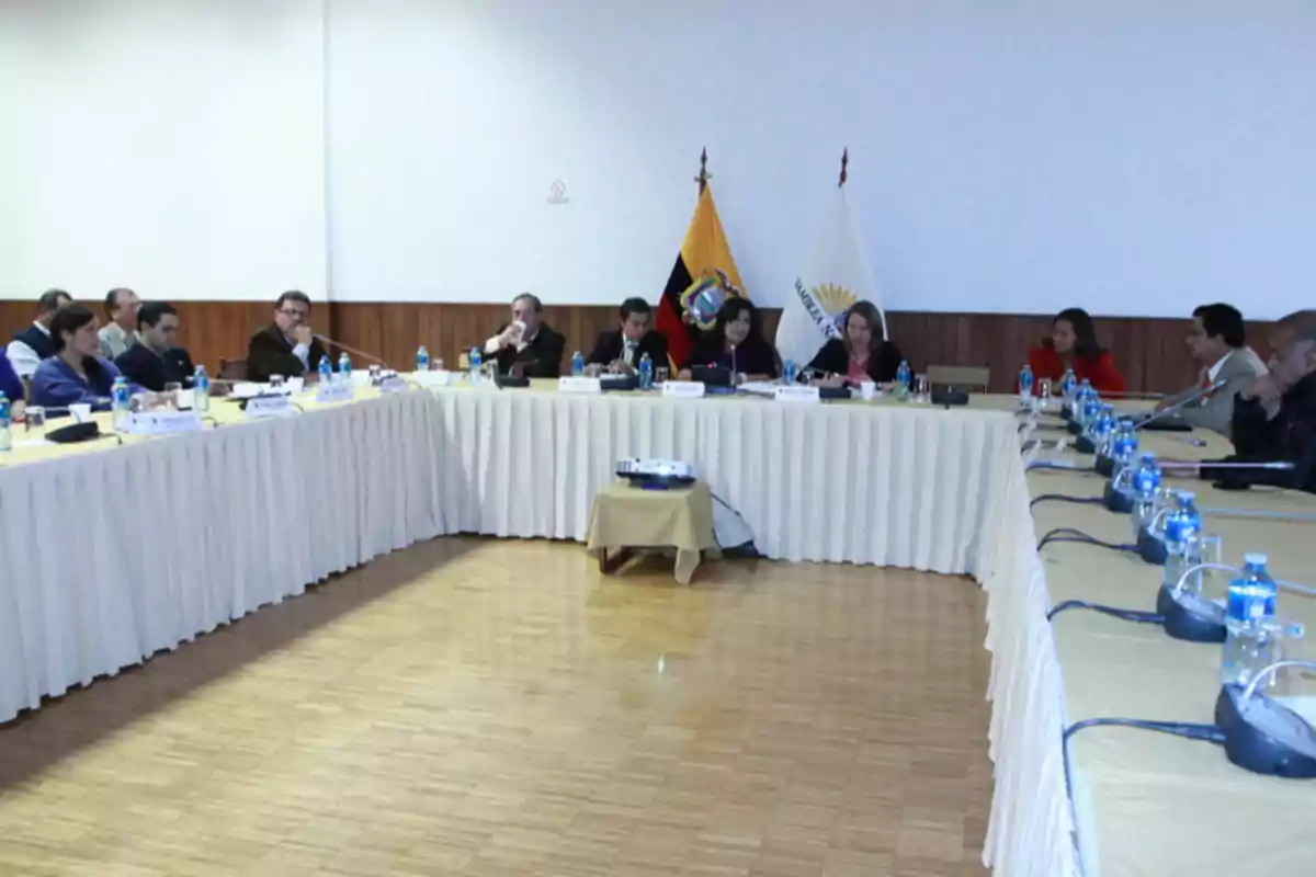 People gathered at a conference table in a room with flags and microphones. People gathered at a conference table in a room with flags and microphones.