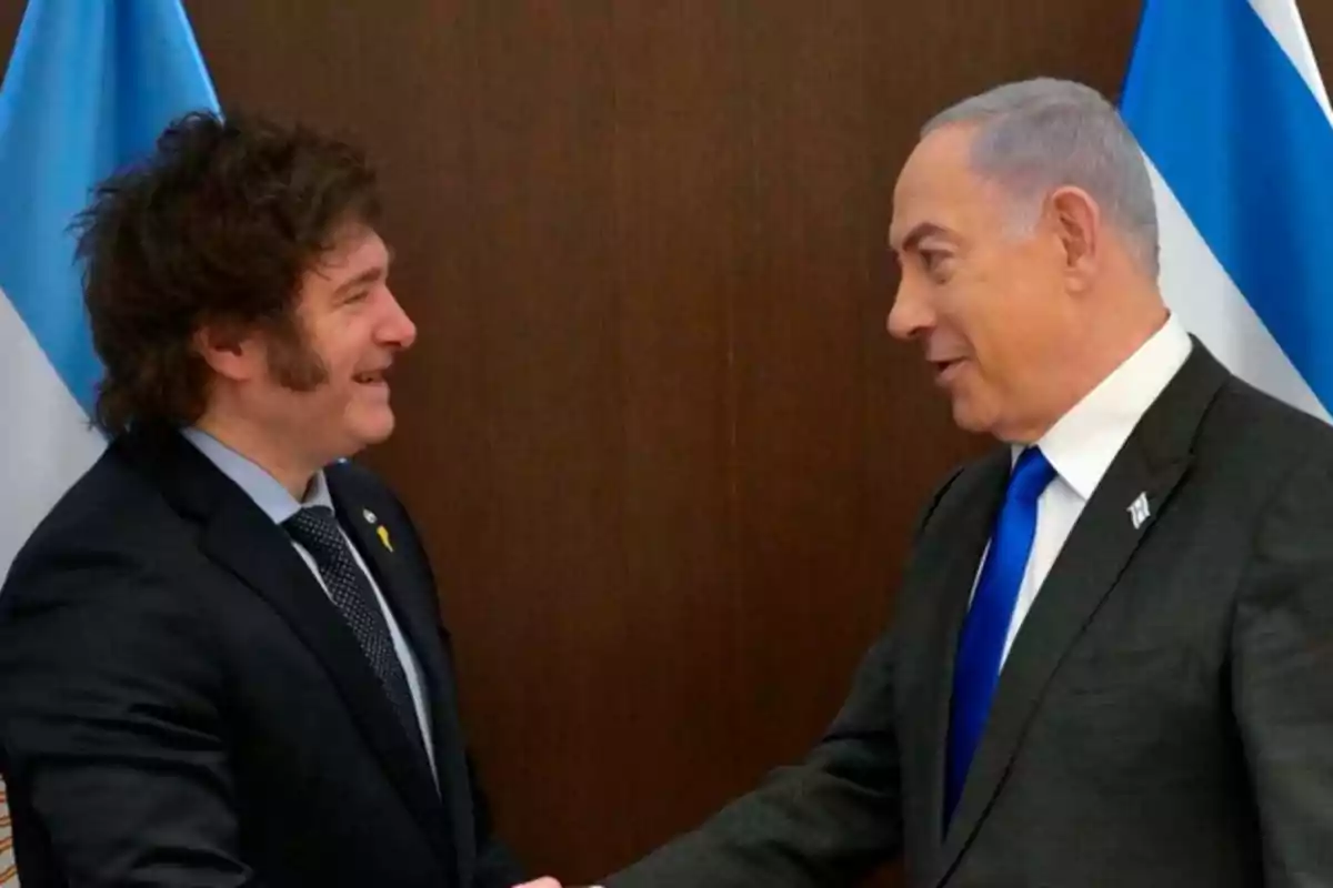 Two men in suits shake hands in front of flags.