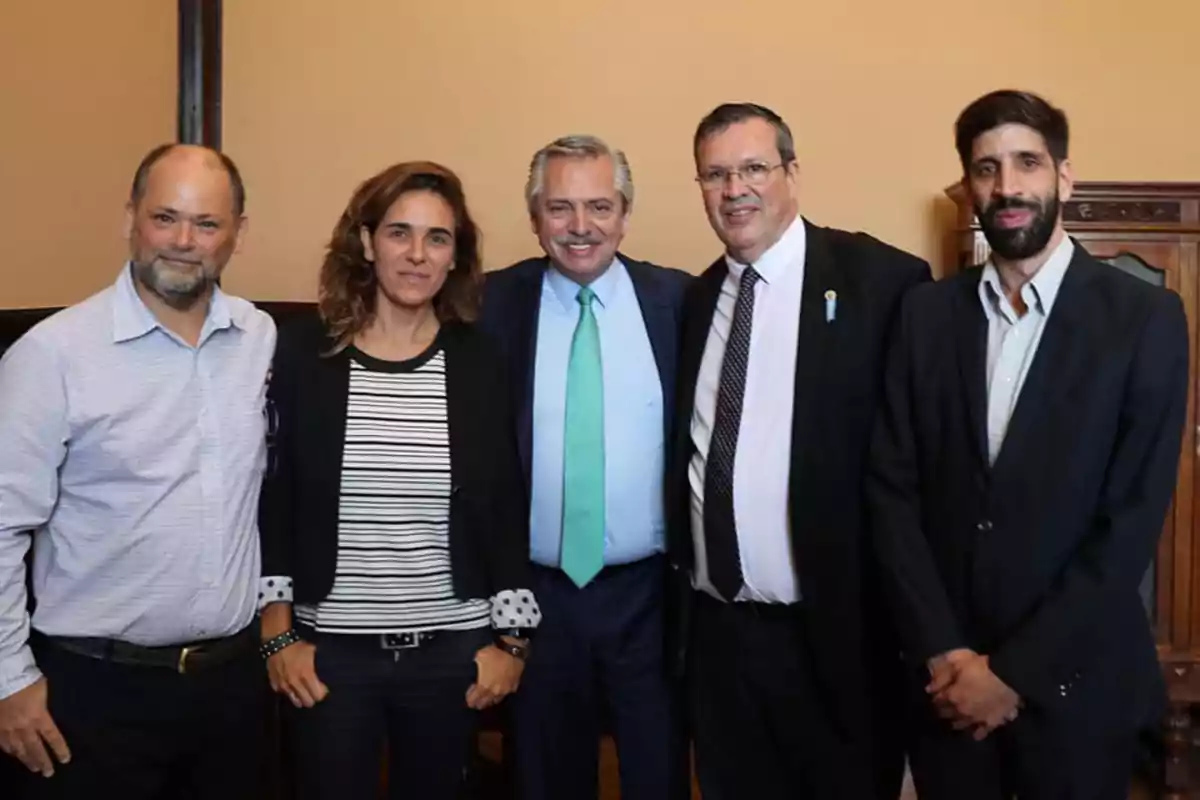 Cinco personas posan juntas en una habitación con paredes de color beige, todos visten ropa formal y sonríen a la cámara.