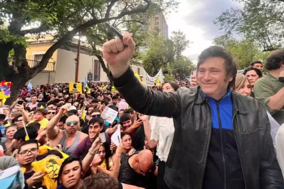 A smiling person in a leather jacket raises his fist in the middle of a crowd during an outdoor demonstration.