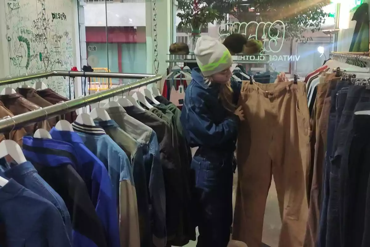 Person with a beanie and denim jacket looking at brown pants in a vintage clothing store. Person with a beanie and denim jacket looking at brown pants in a vintage clothing store.