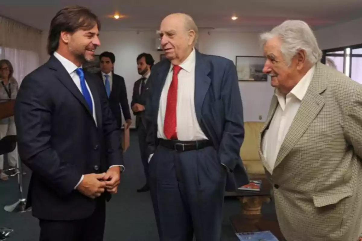 Three men are conversing in a room, two of them are wearing dark suits and the other is wearing a light jacket, while other people are in the background. Three men are conversing in a room, two of them are wearing dark suits and the other is wearing a light jacket, while other people are in the background.