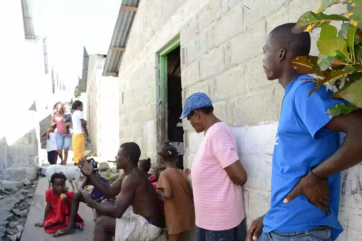 Varias personas de diferentes edades están reunidas junto a una casa de bloques en una calle estrecha y soleada