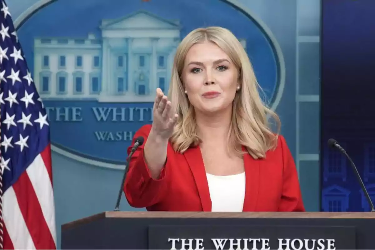 Mujer rubia con saco rojo hablando en un podio frente al logo de la Casa Blanca y una bandera de Estados Unidos Mujer rubia con saco rojo hablando en un podio frente al logo de la Casa Blanca y una bandera de Estados Unidos