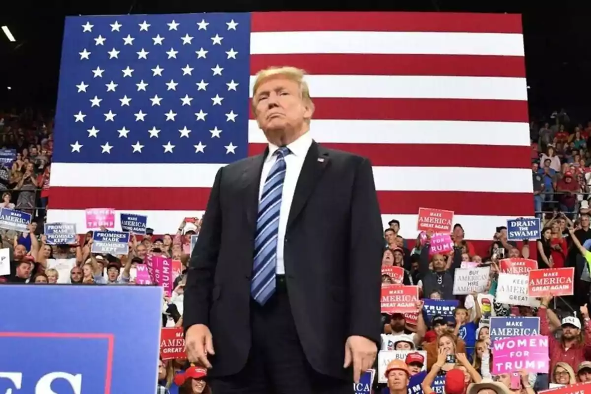 Un hombre de traje frente a una multitud con carteles y una gran bandera de Estados Unidos de fondo.