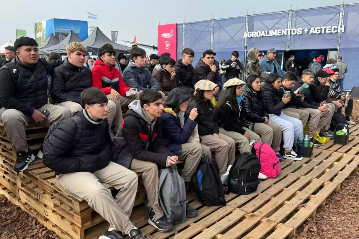A group of young people sitting on bleachers made of wooden pallets attend an outdoor event in front of a booth that says agro-foods agtech. A group of young people sitting on bleachers made of wooden pallets attend an outdoor event in front of a booth that says agro-foods agtech.