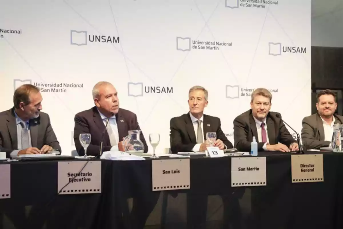 Cinco hombres en traje sentados en una mesa con carteles que indican sus roles, en un evento de la Universidad Nacional de San Martín.