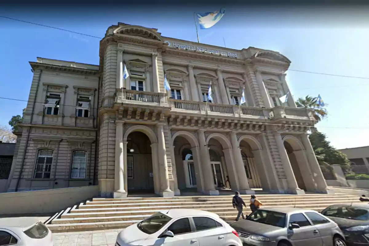 Edificio histórico con columnas y banderas en la fachada, con personas caminando y autos estacionados al frente.