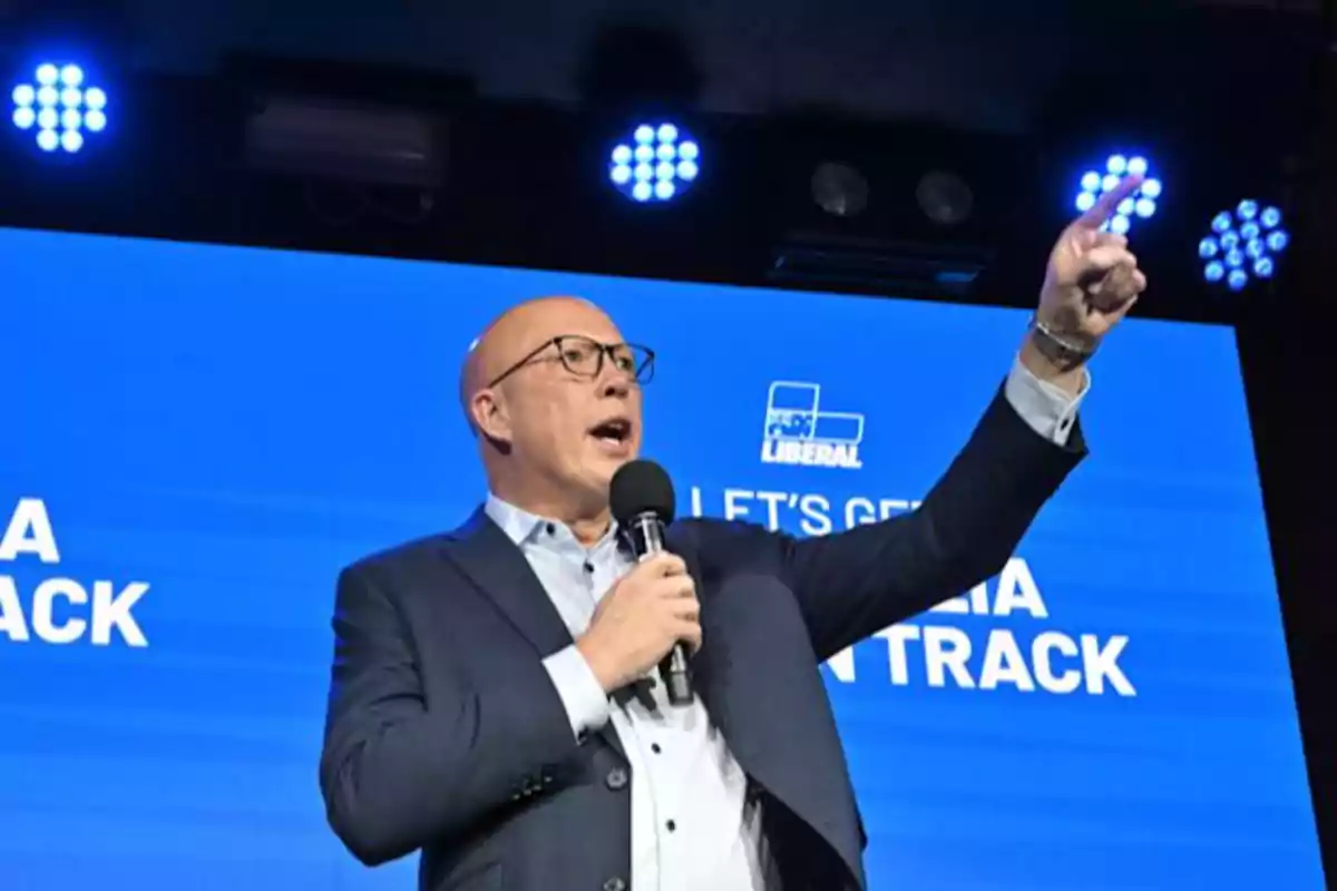 A man with glasses and a microphone speaks on a stage with a blue background and bright lights. A man with glasses and a microphone speaks on a stage with a blue background and bright lights.